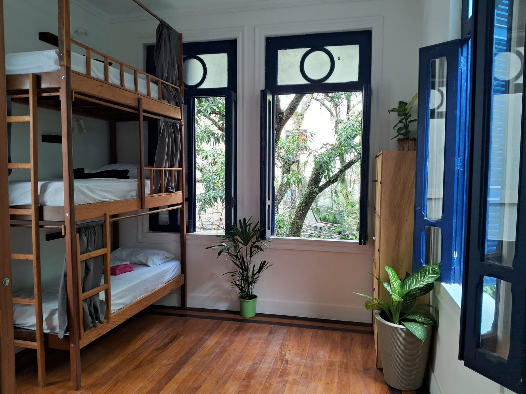 Wooden bunk beds with privacy curtains and tall blue-framed windows in the 9-bed mixed dorm